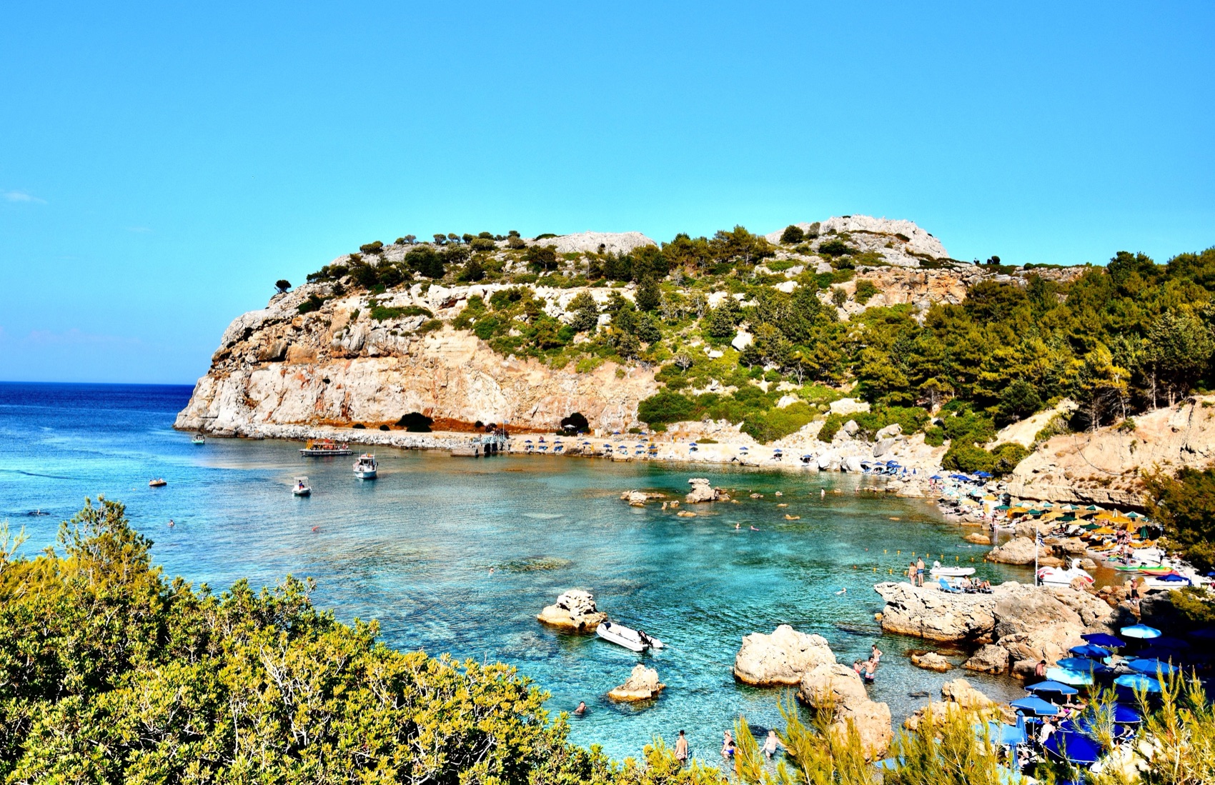 A pine-covered headland above a bay with clear turquoise water and small boats